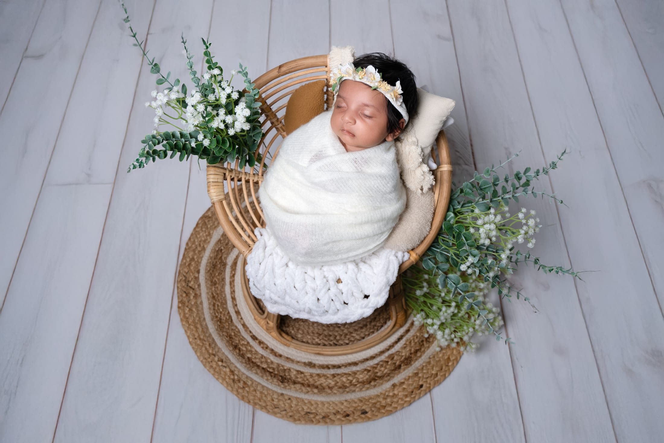 Newborn wrapped in white, resting in a wicker chair with florals